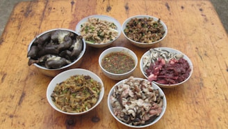Several bowls are arranged on a wooden table, each containing a variety of food items. The bowls include different types of prepared meats, vegetables, and a bowl of sauce with herbs and spices.