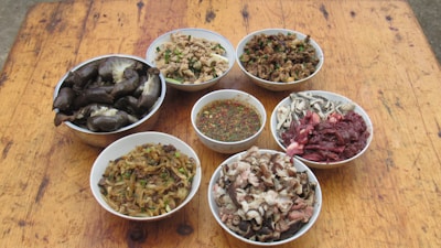 Several bowls are arranged on a wooden table, each containing a variety of food items. The bowls include different types of prepared meats, vegetables, and a bowl of sauce with herbs and spices.