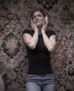 A person wearing headphones lies on an intricately patterned rug, with hands touching the headphones gently. They wear a black top and light blue jeans, appearing relaxed and absorbed in listening.