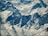 A stunning aerial view of rugged snow-covered mountains with sharp peaks and deep valleys, showcasing the massive, sheer cliffs and intricate patterns formed by the snow and ice.