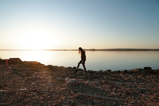 A person is walking along a rocky shoreline with a serene body of water and a bright setting sun in the background. The long shadows and tranquil scene convey a sense of solitude and contemplation.