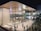 A modern, spacious retail store with large glass windows showcasing the interior, which is bustling with shoppers. The sleek architectural design includes a curved wooden roof and an illuminated logo on the glass facade. People are walking and gathering outside, where a queue setup is present, indicating a possibly busy and popular location.