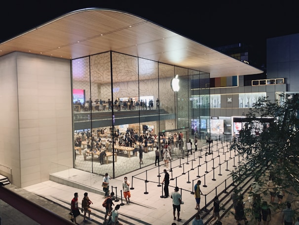 A modern, spacious retail store with large glass windows showcasing the interior, which is bustling with shoppers. The sleek architectural design includes a curved wooden roof and an illuminated logo on the glass facade. People are walking and gathering outside, where a queue setup is present, indicating a possibly busy and popular location.