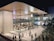 A modern, spacious retail store with large glass windows showcasing the interior, which is bustling with shoppers. The sleek architectural design includes a curved wooden roof and an illuminated logo on the glass facade. People are walking and gathering outside, where a queue setup is present, indicating a possibly busy and popular location.