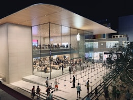 A modern, spacious retail store with large glass windows showcasing the interior, which is bustling with shoppers. The sleek architectural design includes a curved wooden roof and an illuminated logo on the glass facade. People are walking and gathering outside, where a queue setup is present, indicating a possibly busy and popular location.