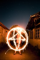 A person stands in a dark alley between two rustic buildings performing light painting with sparklers, creating a glowing intricate pattern that resembles a large pentagram. The night sky is visible with faint stars in the background, adding to the mystical appearance of the scene.