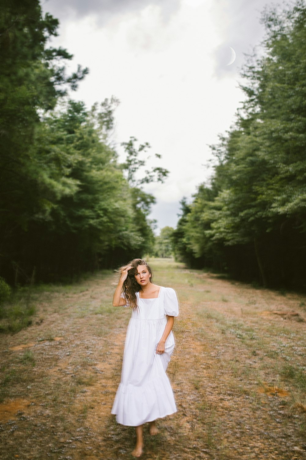 woman in white dress standing on dirt road between green trees during daytime