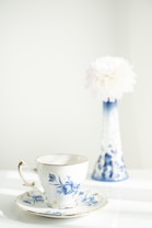 A delicate porcelain cup and saucer with blue floral designs sits in the foreground. In the background, a tall, slender vase with similar blue patterns holds a large, fluffy white flower. The setting is bright and gently lit, suggesting a peaceful and elegant atmosphere.