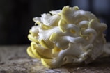 A cluster of yellow and white oyster mushrooms rest on a wooden surface. The mushrooms have delicate, ruffled edges and a smooth texture, with some of the smaller pieces curling inward. The lighting is soft, creating a moody and natural ambiance.