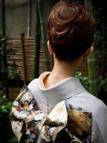 A person is seen from behind wearing a traditional kimono with an intricately designed obi featuring floral patterns. The hair is styled in a neat updo. Bamboo plants and wooden fencing are visible in the background, creating a serene and natural setting.