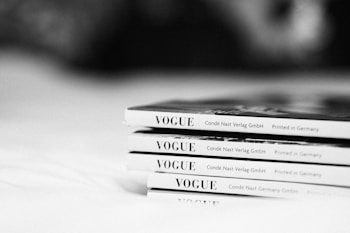 A neatly stacked set of thin magazines with the title 'VOGUE' visible on the spines. The magazines are placed on a soft surface, possibly a bed or a tablecloth. The image is in black and white, giving it a classic and elegant appearance.