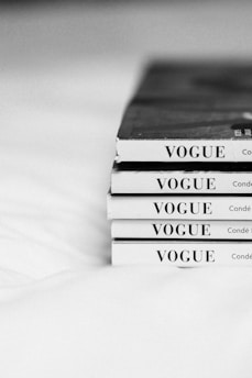 A stack of five Vogue magazines is neatly arranged with their spines facing outward, displaying the magazine title prominently in capital letters. The image is in black and white, giving it a classic and timeless feel. The magazines are placed on a light-colored surface.