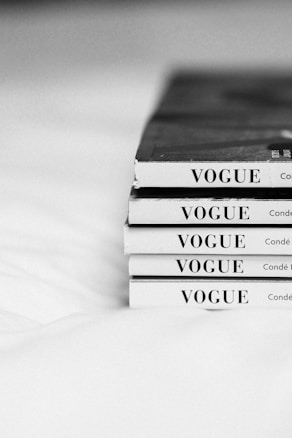 A stack of five Vogue magazines is neatly arranged with their spines facing outward, displaying the magazine title prominently in capital letters. The image is in black and white, giving it a classic and timeless feel. The magazines are placed on a light-colored surface.
