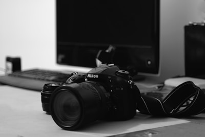 A DSLR camera with a lens, identified as a Nikon, rests on a desk. Behind the camera, a computer monitor, keyboard, and various office items are present, creating an indoor setting with a focus on photography or work-related tasks.