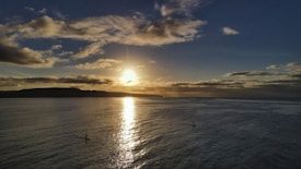 A serene ocean scene at sunset with the sun shining brightly above the horizon, casting a warm glow across the water. Two silhouetted kayakers are peacefully paddling in the calm sea under a sky filled with scattered clouds.