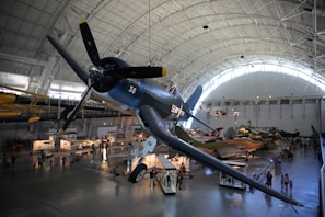 A large indoor aviation museum displays various historic aircraft under a high arched ceiling. A prominent blue fighter plane with the number 56 is suspended in the foreground, surrounded by other aircraft including helicopters and jets. People are walking and observing the exhibits below.