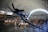A large indoor aviation museum displays various historic aircraft under a high arched ceiling. A prominent blue fighter plane with the number 56 is suspended in the foreground, surrounded by other aircraft including helicopters and jets. People are walking and observing the exhibits below.