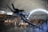 A large indoor aviation museum displays various historic aircraft under a high arched ceiling. A prominent blue fighter plane with the number 56 is suspended in the foreground, surrounded by other aircraft including helicopters and jets. People are walking and observing the exhibits below.