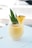 A creamy yellow smoothie or drink is in a clear glass, garnished with a slice of pineapple and green leaves. It is placed on a white wooden surface, with a softly blurred background suggesting an outdoor setting.