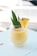 A creamy yellow smoothie or drink is in a clear glass, garnished with a slice of pineapple and green leaves. It is placed on a white wooden surface, with a softly blurred background suggesting an outdoor setting.