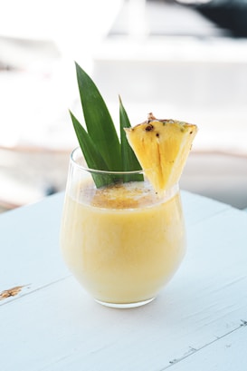 A creamy yellow smoothie or drink is in a clear glass, garnished with a slice of pineapple and green leaves. It is placed on a white wooden surface, with a softly blurred background suggesting an outdoor setting.