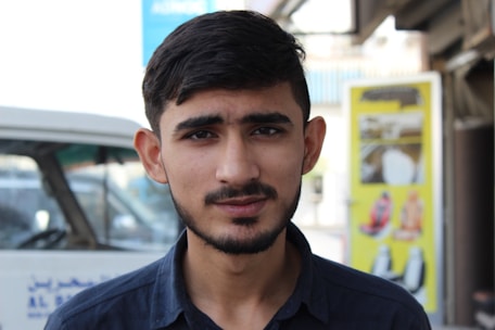 A young man with short dark hair and a beard is standing in an outdoor setting. He is wearing a dark shirt and looking directly at the camera. Behind him, there is a partially visible white vehicle with some text in another language. To the right, a yellow advertisement poster is seen, featuring various images.