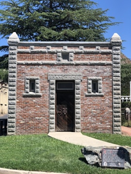 A small, historical brick building with a rustic wooden door, labeled 'Branch Jail' above the entrance. The building has stone accents along the edges and corners, with decorative carving details. It is set against a backdrop of lush greenery, with a prominent large tree. A stone plaque is positioned on the grassy area in front of the building.
