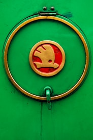A circular emblem with a gold outline and red interior, featuring a stylized symbol, is displayed on a vibrant green background. A handle extends from the lower part of the circle, indicating a functional aspect.
