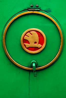 A circular emblem with a gold outline and red interior, featuring a stylized symbol, is displayed on a vibrant green background. A handle extends from the lower part of the circle, indicating a functional aspect.