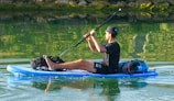 A person is seated on an inflatable paddleboard, paddling through calm water. The individual is dressed casually, wearing a black t-shirt, shorts, and a cap, carrying backpacks and gear on the paddleboard. The water reflects greenery from the surrounding vegetation.