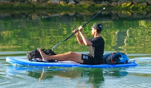 A person is seated on an inflatable paddleboard, paddling through calm water. The individual is dressed casually, wearing a black t-shirt, shorts, and a cap, carrying backpacks and gear on the paddleboard. The water reflects greenery from the surrounding vegetation.