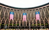 An architectural facade with intricate geometric patterns and large glass windows features three prominently displayed Malaysian flags. Several palm trees are lined up in front of the structure, adding a tropical element to the scene.