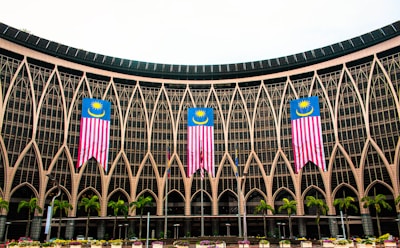 An architectural facade with intricate geometric patterns and large glass windows features three prominently displayed Malaysian flags. Several palm trees are lined up in front of the structure, adding a tropical element to the scene.