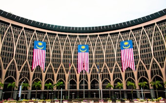 An architectural facade with intricate geometric patterns and large glass windows features three prominently displayed Malaysian flags. Several palm trees are lined up in front of the structure, adding a tropical element to the scene.