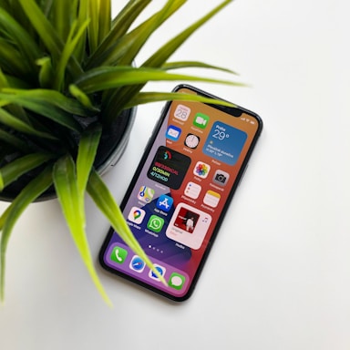 A smartphone displaying various colorful app icons on its screen rests on a white surface next to a green potted plant. The device is lit up, showing clear and vibrant digital content, including weather information and calendar apps.