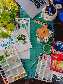 A creative workspace is filled with various art supplies including watercolors, brushes, colored pencils, and a painting of flowers. A laptop is partially visible at the top, and there's a ceramic holder in the shape of an elephant containing pens and brushes. A green leafy plant adds a natural element to the scene.