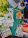 A creative workspace is filled with various art supplies including watercolors, brushes, colored pencils, and a painting of flowers. A laptop is partially visible at the top, and there's a ceramic holder in the shape of an elephant containing pens and brushes. A green leafy plant adds a natural element to the scene.