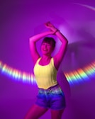 A person stands joyfully with arms raised against a vibrant purple background, creating a playful and energetic atmosphere. Light reflects around them in a colorful arc, casting hues of blue, yellow, and pink. The person is wearing a bright yellow top and denim shorts, adding to the lively scene.