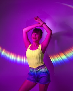 A person stands joyfully with arms raised against a vibrant purple background, creating a playful and energetic atmosphere. Light reflects around them in a colorful arc, casting hues of blue, yellow, and pink. The person is wearing a bright yellow top and denim shorts, adding to the lively scene.