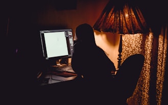 A silhouette of a person sitting at a desk using a computer in a dimly lit room. The room is illuminated by a warm, patterned lamp and partially visible curtains. The computer screen displays a drawing or graphic design software.