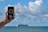 A hand is holding a smartphone with a compass app displayed on the screen. The background features a large cruise ship sailing on the ocean under a partly cloudy sky. The phone shows a compass reading of 201° S, along with location details indicating Bournemouth, England.