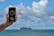 A hand is holding a smartphone with a compass app displayed on the screen. The background features a large cruise ship sailing on the ocean under a partly cloudy sky. The phone shows a compass reading of 201° S, along with location details indicating Bournemouth, England.