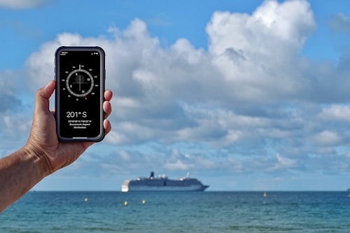A hand is holding a smartphone with a compass app displayed on the screen. The background features a large cruise ship sailing on the ocean under a partly cloudy sky. The phone shows a compass reading of 201&deg; S, along with location details indicating Bournemouth, England.
