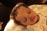 A peacefully sleeping baby with curly hair rests on a cushion decorated with cartoon elephant patterns. The soft lighting casts a warm, golden glow over the scene.