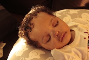 A peacefully sleeping baby with curly hair rests on a cushion decorated with cartoon elephant patterns. The soft lighting casts a warm, golden glow over the scene.