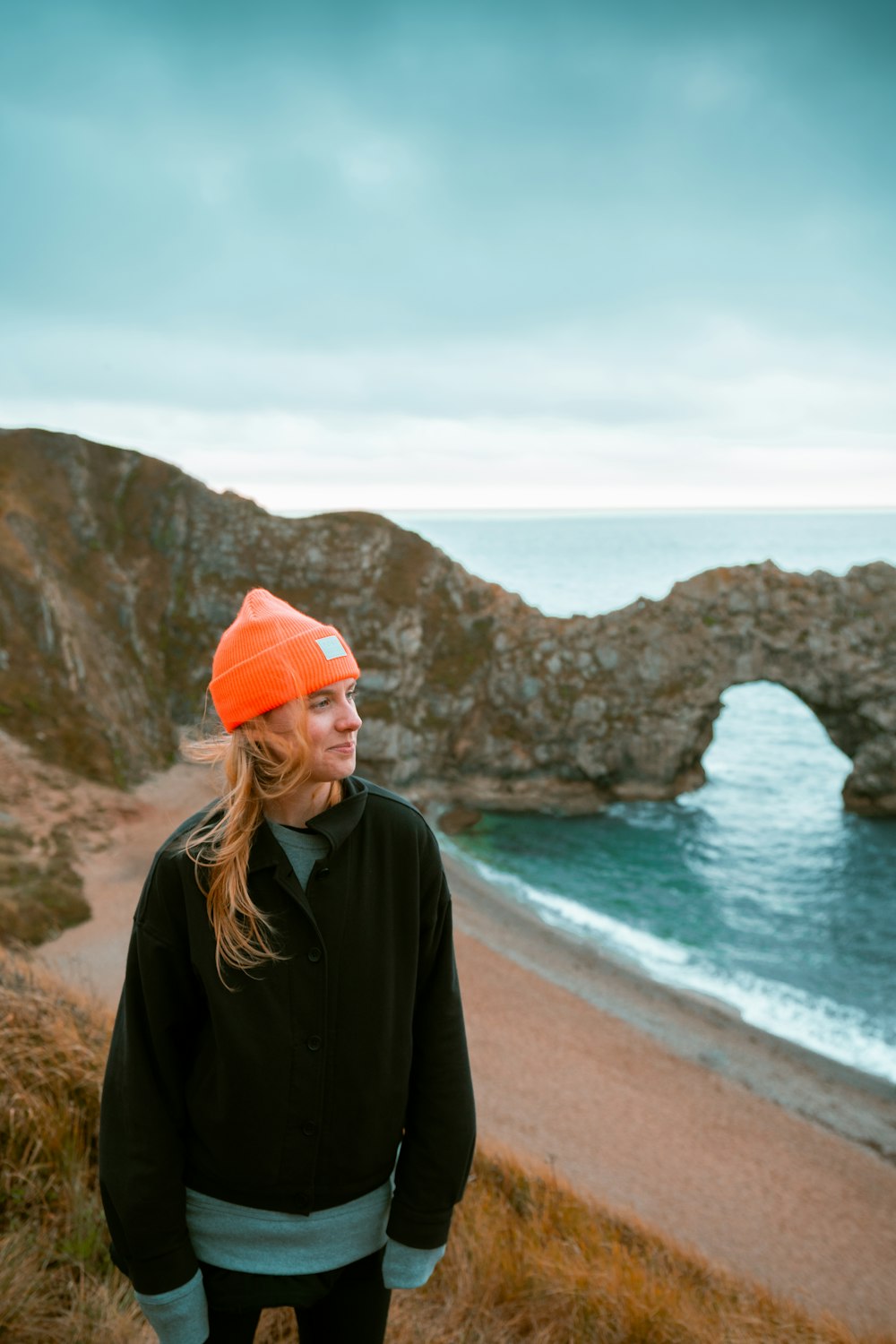 woman in black jacket and orange knit cap standing on beach shore during daytime