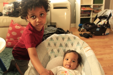 Two young children are in a warmly lit room. One child, standing, wears a red shirt and looks directly at the camera with a serious expression. The other child is lying in a patterned baby seat, smiling playfully with their tongue out.