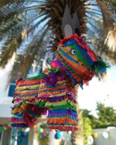 A colorful, striped pi&ntilde;ata shaped like a donkey is suspended in front of a palm tree in a sunny outdoor setting. The pi&ntilde;ata features a pattern of vibrant paper streamers in shades of red, blue, green, yellow, orange, and purple, creating a festive appearance. The background consists of palm leaves and blurred elements, suggesting a party or celebration atmosphere.