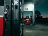A gas station at twilight with visible fuel pumps displaying the numbers '95' and 'E10'. The station is dimly lit, with a dark background showing silhouettes of trees against a dusky sky. The structures are metallic and red, and there are Cyrillic letters on the station signs.