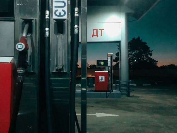 A gas station at twilight with visible fuel pumps displaying the numbers '95' and 'E10'. The station is dimly lit, with a dark background showing silhouettes of trees against a dusky sky. The structures are metallic and red, and there are Cyrillic letters on the station signs.